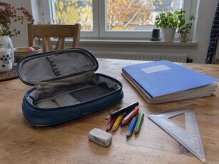 Ausgeräumtes Schulmäppchen mit kurzen Buntstiften, Radiergummi, Geodreieck und Heften auf einem Holztisch